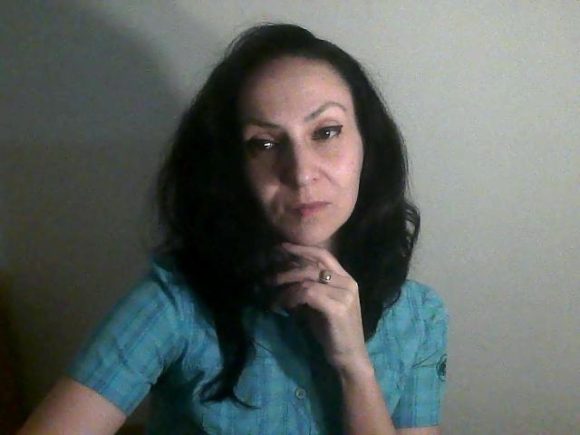 UrbanKate with dark, wavy hair, wearing a blue shirt, has a serious face, holding her chin with a ringed finger, against a plain background.