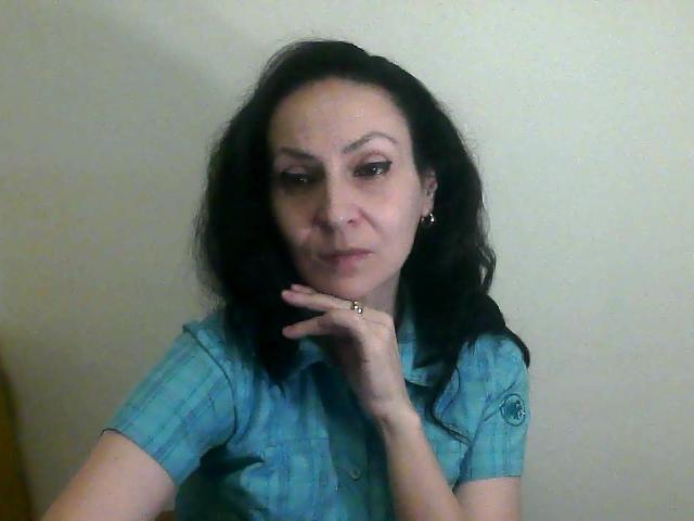 UrbanKate with dark wavy hair, wearing a blue plaid shirt, has her chin on her hand, showing a ring on her finger, and a slightly smug expression.