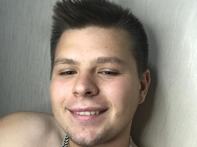MagicH's shirtless face with short, spiked brown hair, slight beard, smiling, and a chain around his neck. His eyes are half-open, and he's leaning against a white wall.