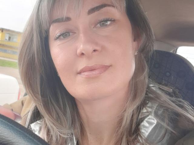 JuliaaCherry, com olhos cinzentos, cabelo castanho claro e maquiagem discreta, sentada em um carro, vestindo uma camisa prateada, olhando para frente com um leve sorriso. Seus seios estão parcialmente visíveis..
