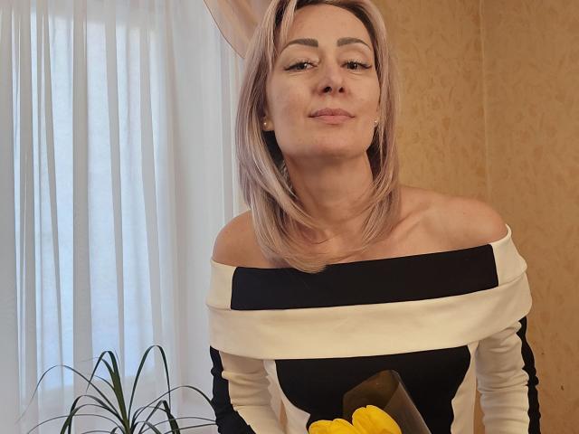 JuliaaCherry with blonde hair, off-shoulder black and white top, holding yellow lemons, smiling, slightly tilting head, white curtains in background, small green plant on left.