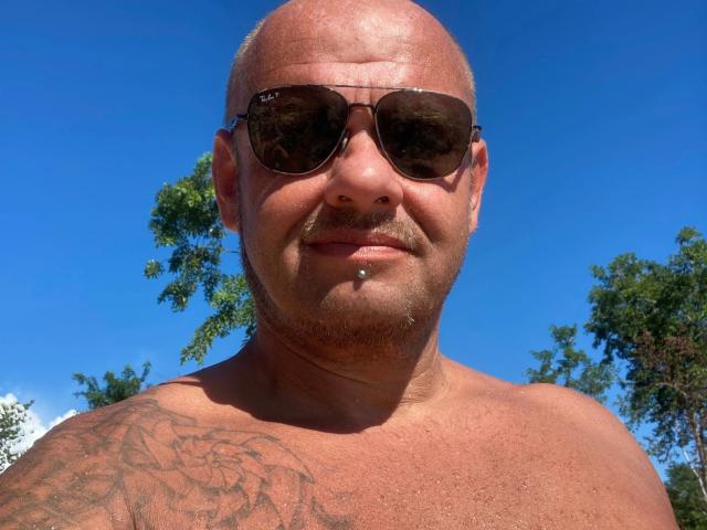 Magic-Mike-81-hot, bald with sunglasses, shirtless, tattoo on right shoulder, slight beard, mustache, nose piercing, blue sky, trees in background, smiling, sunlight.