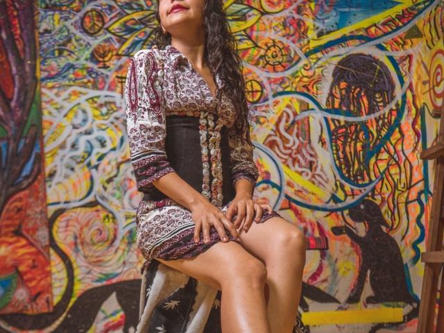 SweetyAloe with curly hair, wearing a patterned dress, sitting on a stool, legs together, hands on knees, against a colorful, abstract, graffiti-style wall. Boobs, ass, pussy not visible.