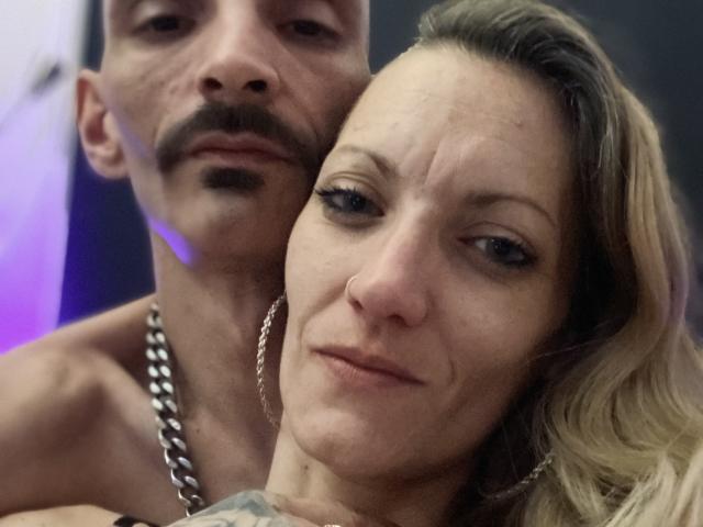 DaneeetJoe's face is in the foreground, showing her nose and lips, with her blonde hair. DaneeetJoe's shirtless, mustached, and chain-necked in the background, slightly out of focus.