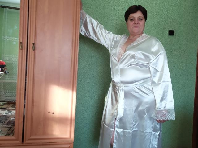 ElzaJewel is a chubby woman with short black hair, wearing a shiny white silk robe, standing with one arm against a wooden wardrobe. Her tits are visible.