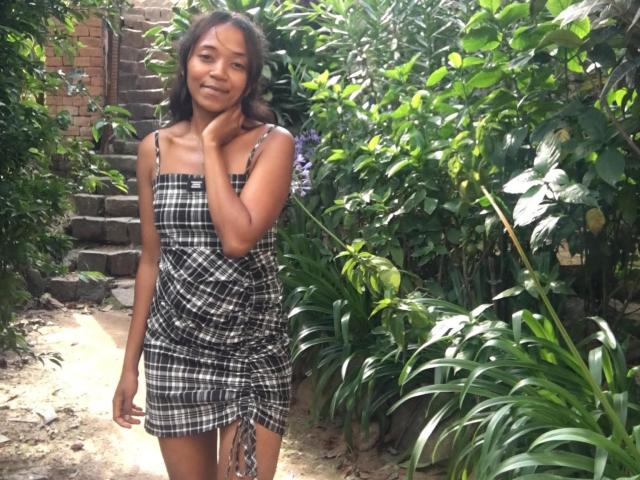 Chattye with small tits, wearing a black and white checkered dress, standing in a garden, smiling, hand on cheek, green plants, and a brick wall in the background.