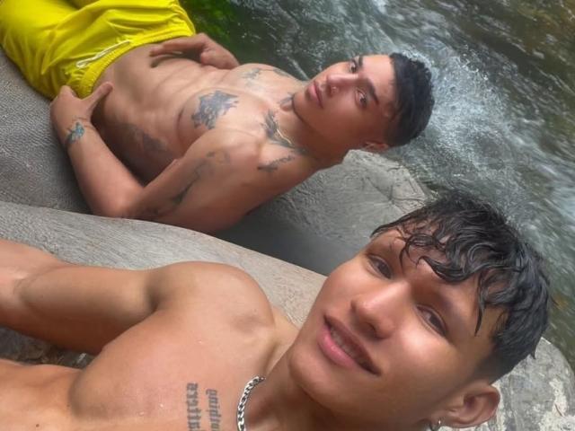 AdonisXAnubis con cabello negro mojado, tatuajes pequeños y un collar, sin camisa, sonriendo en primer plano. AdonisXAnubis con pantalones cortos amarillos, tatuajes y pecho mojado, acostado en el fondo..