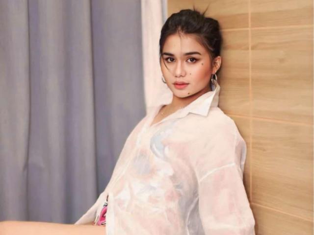 AndyWalton with moles on her cheeks, wearing a sheer white shirt, black hair in a bun, hoop earrings, leaning against a wooden wall, looking at the camera.