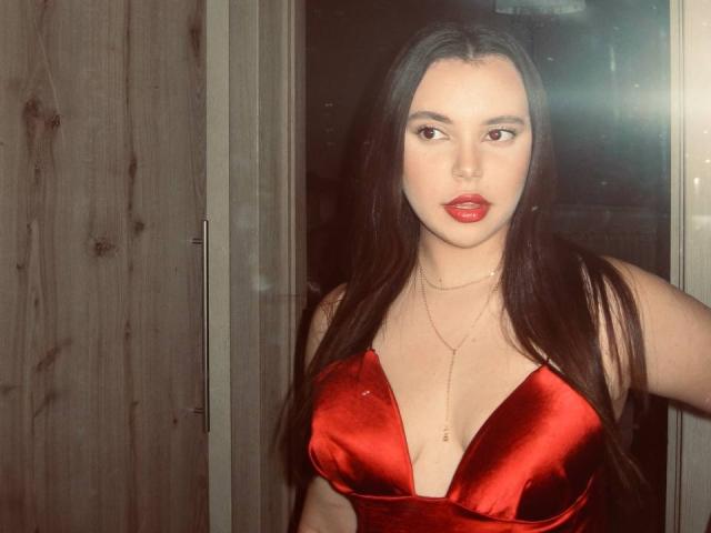 JennaKittens with long dark hair, red satin dress, deep cleavage, red lips, gold necklace, wooden door, and a slightly seductive look. Boobs are prominent, tits are noticeable.