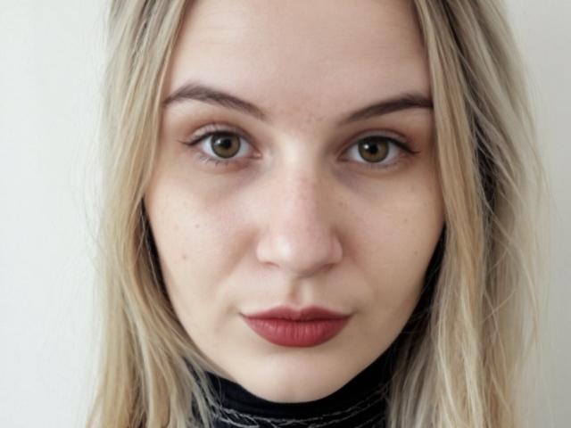 KatrinaPetrova's face with blonde hair, green eyes, and red lipstick, wearing a black turtleneck, looking straight at the camera with a subtle smile.