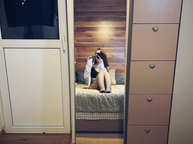 KatrinaPetrova with long brown hair, wearing fishnet stockings and black heels, sitting on a bed, holding a white shirt, reflected in a mirror, in a wooden-walled room.