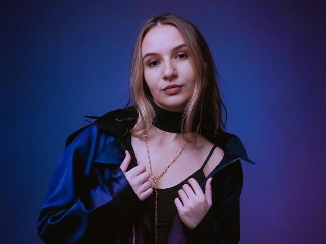 KatrinaPetrova with light brown hair, wearing a black choker, blue jacket, and gold necklace, holding jacket edges, purple background, slight smile, looking at camera.