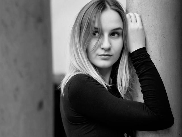 KatrinaPetrova with blonde hair, wearing a black long-sleeve shirt, leaning against a column, looking slightly to the side, hand on column, in a black-and-white photo.