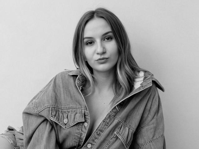 KatrinaPetrova with medium tits, wearing an unbuttoned denim jacket, has a pouty face, and wavy, shoulder-length hair in a black-and-white photo.