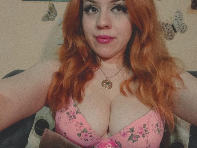 SpicyGrace con i capelli arancioni, reggiseno floreale rosa, che mostra un seno generoso, rossetto rosa, collana dorata, sorridente alla telecamera, che tiene una coperta marrone..