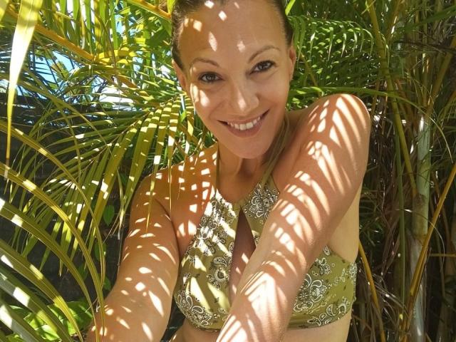 LylyFrench with smiling face, brown eyes, and green lace-up bikini top, standing among sunlit palm leaves, casting shadow patterns on her skin. She has a cute, attractive look.