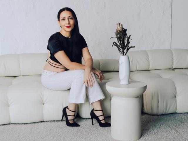 LiluGrants with dark hair, red lipstick, black top, white pants, black strappy heels, sitting on white sofa, hands on knees, white circular table with vase of dried flowers.