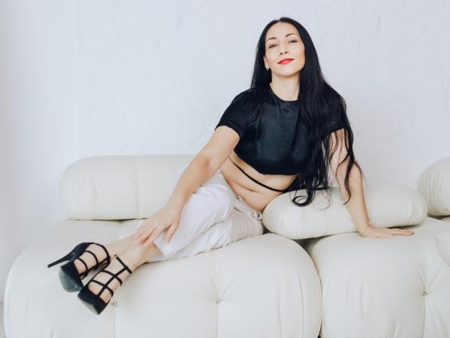 LiluGrants with long black hair, red lipstick, black crop top, white pants, black strappy heels, sitting on white leather couch, showing her flat stomach and sexy legs.