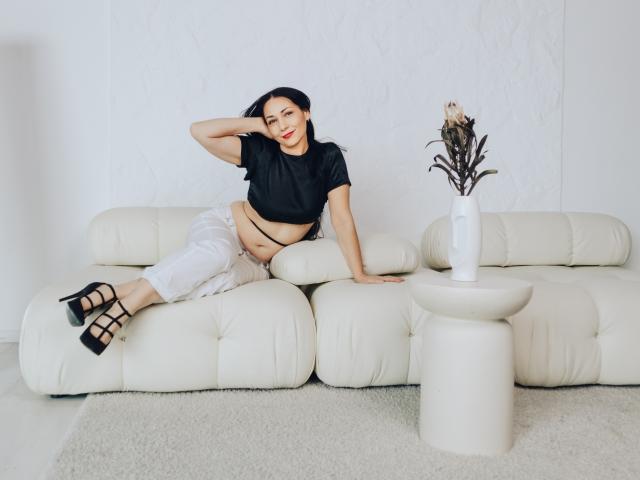 LiluGrants with dark hair, red lipstick, black crop top, white pants, black strappy heels, lying on white leather sofa, hand in hair, white table with vase in foreground.