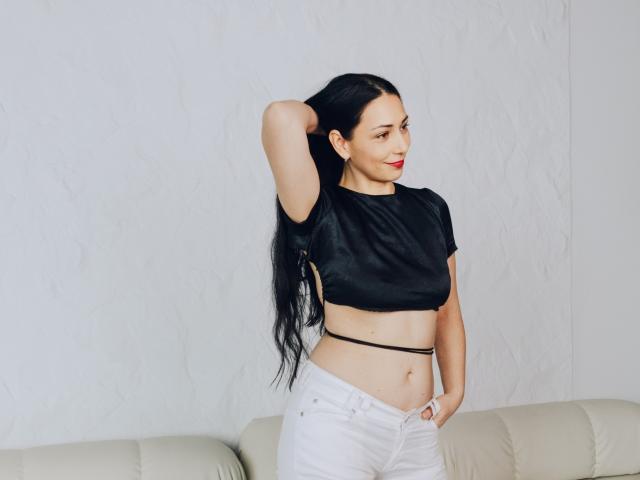 Lilu, con cabello negro largo, lápiz labial rojo, top corto negro, pantalones blancos y cinturón negro, de pie con un brazo detrás de la cabeza, sonriendo, con una mano en el bolsillo, sobre un sofá beige..