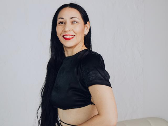 Lilu, con i suoi lunghi capelli neri, il rossetto rosso, la canotta nera e il sorriso, mostra il seno contro un muro bianco, e ha un aspetto carino, sexy e attraente..