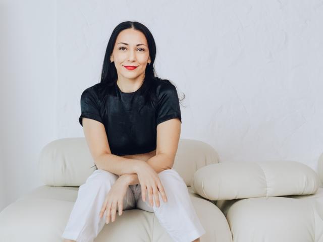 LiluGrants, sitting on a white leather couch, wearing a black shirt and white pants, with hands on knees, smiling, red lipstick, black hair, light skin.