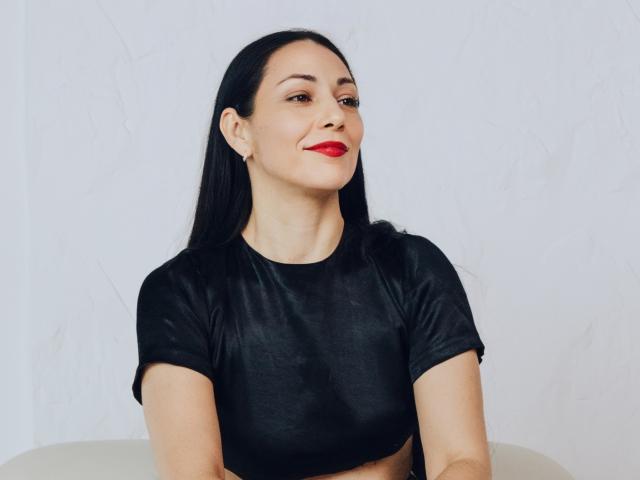 LiluGrants has dark hair, red lipstick, and a slight smile. She's wearing a black shirt, arms crossed, and looking to the side. Her ass is not visible.