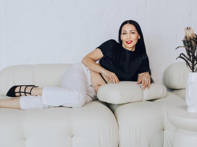 LiluGrants with black hair, red lipstick, black shirt, white pants, and black heels, lying on a white leather couch, exposing her flat belly and black underwear.