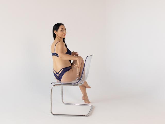 LiluGrants, Asian woman with long black hair, wearing navy blue lace bra and panties, sitting on a clear acrylic chair, legs crossed, bare feet, looking to the side.