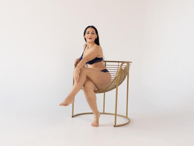 LiluGrants sitting on a gold chair, wearing a navy blue bikini, showing her tits and ass, legs crossed, smiling, barefoot, with long black hair, in a white room.