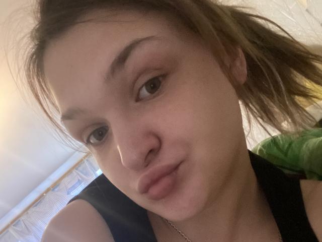BabeKissAli with puffy lips, light brown eyes, and slightly wavy brown hair in a ponytail, wearing a black top, looking at the camera with a small smile.
