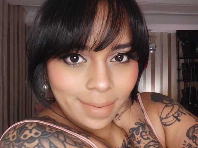 Chili con i capelli neri e tatuaggi sta dando un footjob, occhi spalancati, sorridente, indossando spaghetti rosa, al chiuso, illuminato da luci a soffitto.