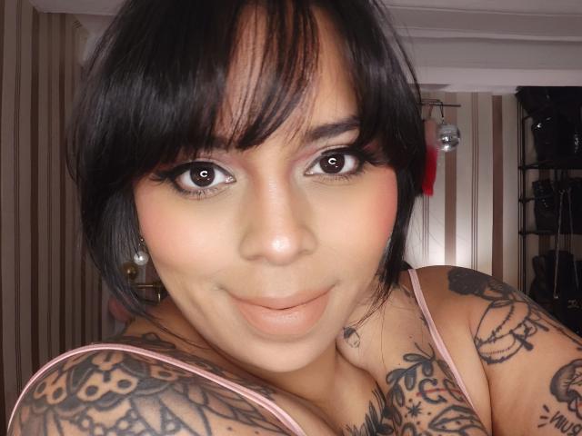 Chili, a sexy Latina woman with black hair and bangs, has large brown eyes, and is smiling while showing off her tattooed shoulders.