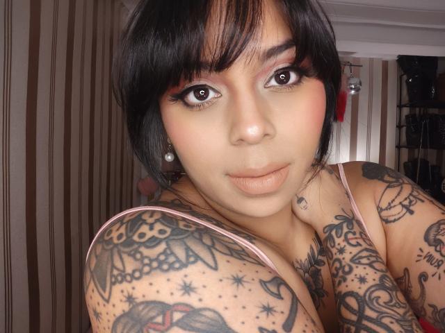 Chili, a heavily tattooed bimbo with black hair, large tits, and a pink tank top, gazes directly at the camera.