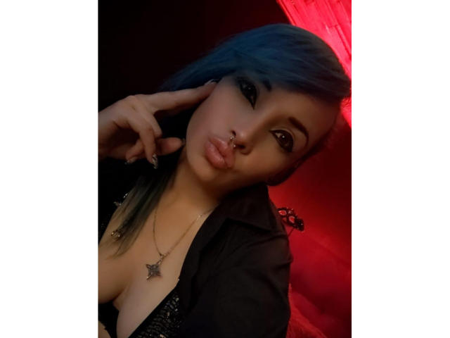 DalilaMusa with blue hair, pouty lips, nose ring, black shirt, and star necklace, pointing at her ear with a finger. Her tits are slightly visible. Red light in background.