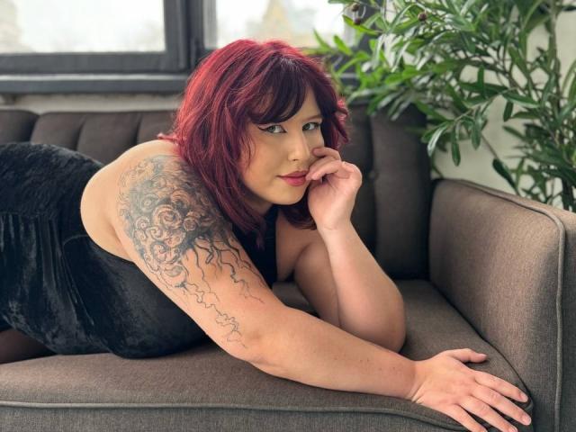 LiliyaMoon met rood haar, een zwarte fluwelen jurk en een getatoeëerde arm, liggend op een grijze bank, haar kin rustend op haar hand, kijkend naar de camera, met een plant in de buurt..