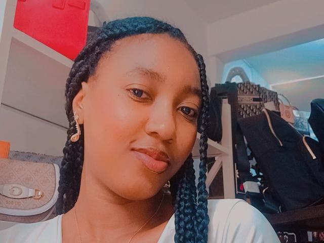 Mamashala with dark skin, blue braided hair, small gold earrings, and a slight pout, wearing a white shirt, in a room with bags and a red object.