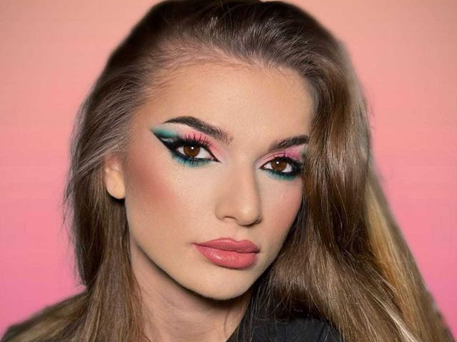 NovaIngrid69 has long brown hair, pink lipstick, and dramatic teal and pink eyeliner. Her brown eyes are slightly upturned, giving a sultry look. Her tits are small, unobscured.
