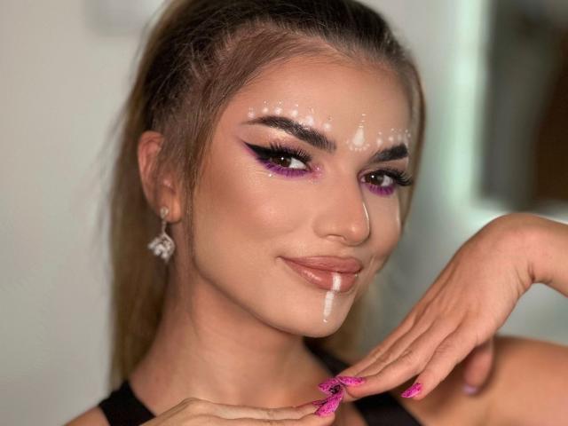 NovaIngrid69 ha il viso pieno di sperma, con ombretto viola, unghie rosa e un sorriso. Indossa una maglietta nera e ha un piercing al naso, con i capelli intrecciati..