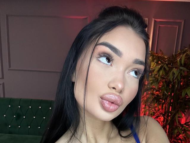SashaFerrera with glossy pink lips, dark eyebrows, long black hair, and blue eyes, looking to the side, wearing a blue top, in a well-lit indoor setting.