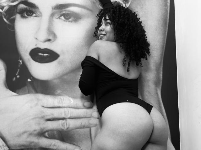 RebeccaMils, avec ses cheveux bouclés, un haut noir à une épaule et un string, montrant ses fesses, se tient devant un grand portrait noir et blanc d'elle-même, avec un rouge à lèvres foncé..