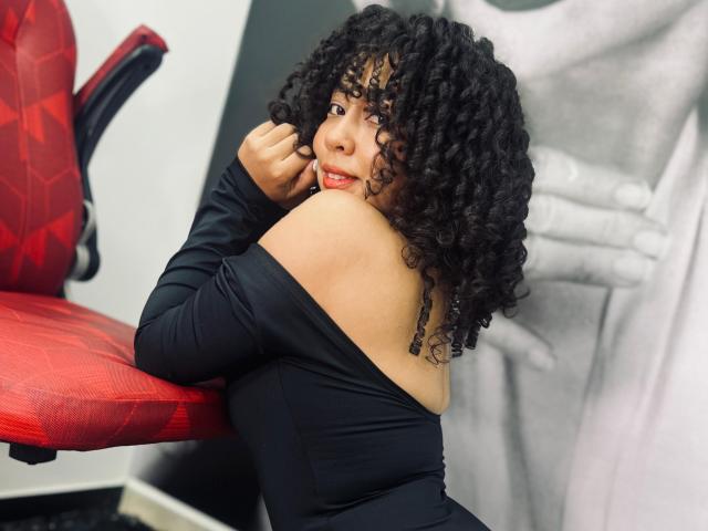 RebeccaMils, con i capelli neri ricci, indossa una maglietta nera con una spalla scoperta, è appoggiata a una sedia rossa, guarda oltre la spalla con un leggero sorriso, mostrando la sua spalla nuda..