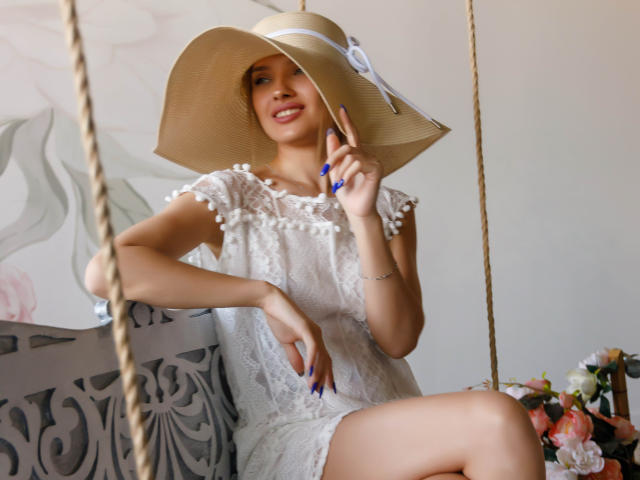 SheillaAdrian sitting on a swing, wearing a white lace dress and a wide-brimmed beige hat, showing blue nails, smiling, with a hand near her face.