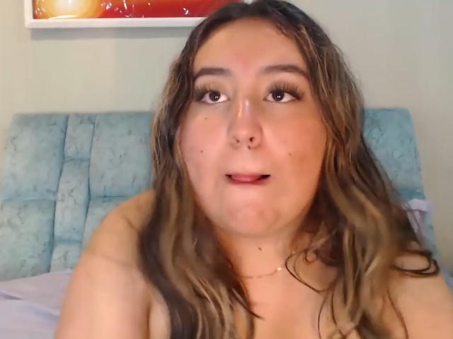 SalomettX with wavy brown hair, nose ring, and long eyelashes, looking up with a slight smile, bare shoulders, and a subtle necklace, in a casual selfie.