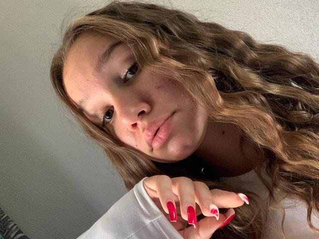 MisaQueen met golvend bruin haar, lichte huid en rode nagels, die lichtjes haar lipjes bijt met haar hand in de buurt van haar gezicht. Ze draagt een wit shirt en ziet er schattig en ontspannen uit..