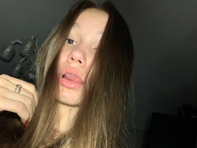 MisaQueen heeft lang bruin haar, lichtjes open lippen met haar tong uitgestoken, en ze steekt haar middelvinger op. Ze draagt een ring aan haar rechterhand..