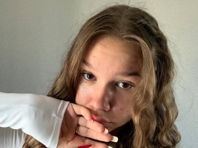 MisaQueen with wavy brown hair, brown eyes, and a slight smirk, touching her nose with a finger, wearing a white long-sleeve shirt, showing red and white nails.