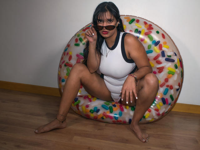 Maria Smith, con senos grandes, vestida con un traje de baño blanco de una sola pieza, está en cuclillas sobre un flotador colorido, usando gafas de sol y lápiz labial rojo, sobre un piso de madera, con piel morena y cabello negro..