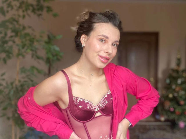 MoonriseGoddessx-hot with dark brown hair in a messy bun, pink cardigan off-shoulder, maroon lace bra with white embroidery, hands on hips, smiling, pink lipstick, small earrings.