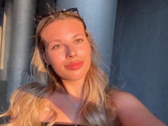MoonriseGoddessx-hot with blonde hair, blue eyes, and red lipstick, wearing a necklace, sunglasses on head, sunlight highlighting her face, showing her tits slightly, in a relaxed pose.